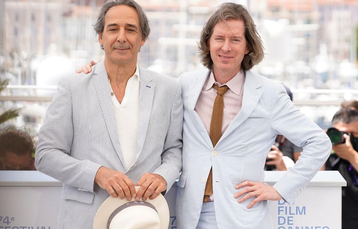 Two men in light striped suits pose together at Cannes 2021, one holding a white hat; photographers and harbor backdrop in the background.