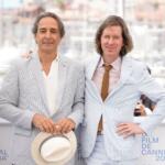 Two men in light striped suits pose together at Cannes 2021, one holding a white hat; photographers and harbor backdrop in the background.