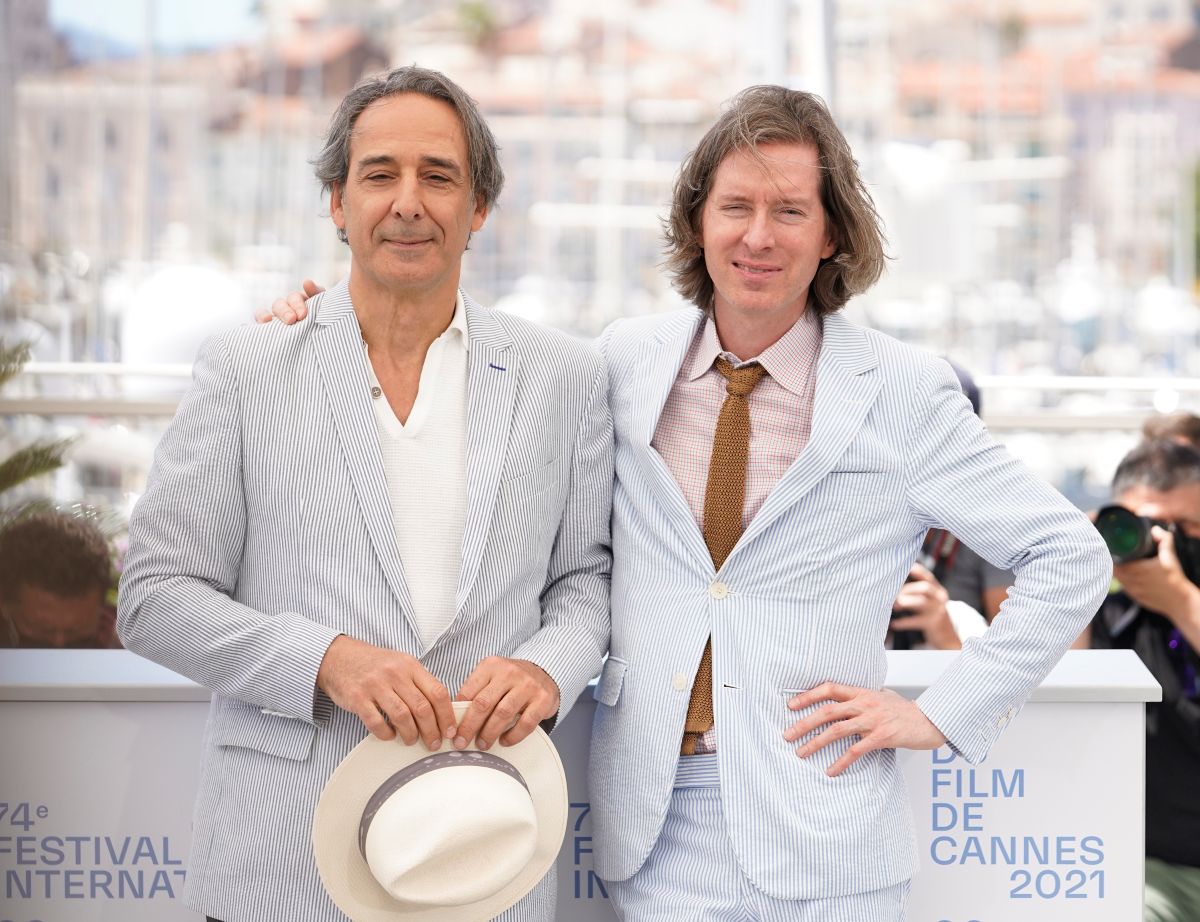 Two men in light striped suits pose together at Cannes 2021, one holding a white hat; photographers and harbor backdrop in the background.