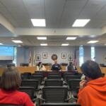 Panel-style meeting room with people seated at a long desk, projector screen reading 'URBANA COMMUNITY RESPONDERS: INPUT ON KEY DECISION POINTS' on the left, and framed portraits and flags on the wall behind.