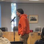Side view of a man in an orange hoodie speaking into a microphone at a podium in a conference room, with audience members in the foreground.