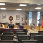 A man in an orange hoodie speaks at a microphone in a formal council chamber, with officials seated at a long desk and framed portraits on the wall behind them.