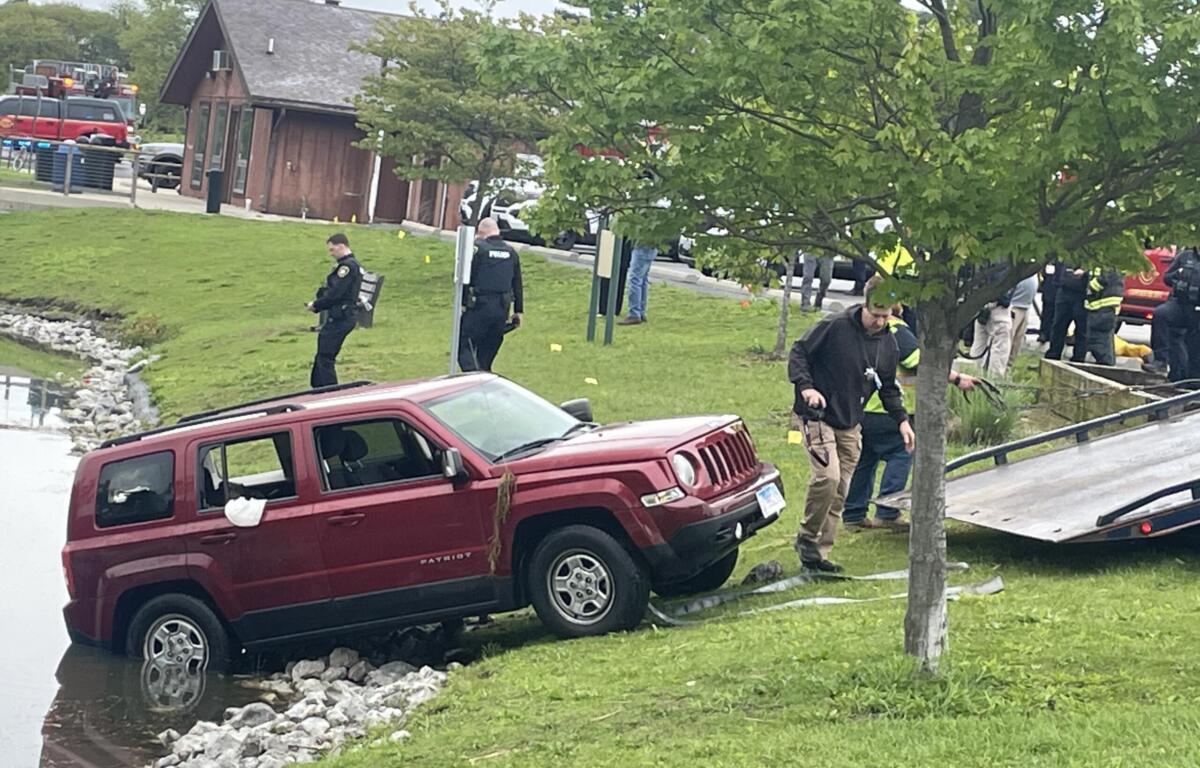 Red Jeep partially in water along a rocky riverbank with emergency responders and bystanders nearby.