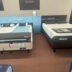 Showroom with several white mattresses on display and blue promotional panels along the wall.