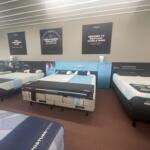 Showroom with multiple Tempur-Pedic mattresses on display and large promotional posters on the wall behind them.