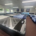 Showroom filled with rows of mattresses on bed frames, with pillows along the side and bright overhead lighting.