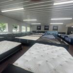 Rows of mattresses on display in a bright showroom with large windows on the left side.