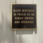 Gold-letter plaque mounted on a beige wall reading: 'ILLINI MATTRESS IS PROUD TO BE FAMILY OWNED AND OPERATED'