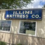 Blue storefront sign reading 'ILLINI MATTRESS CO.' on a beige building, with an open neon sign in the window and trees above.