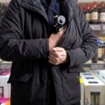Person in a black jacket stands in a liquor store aisle, arms crossed and holding a bottle with a small metal device, shelves of bottles in the background.