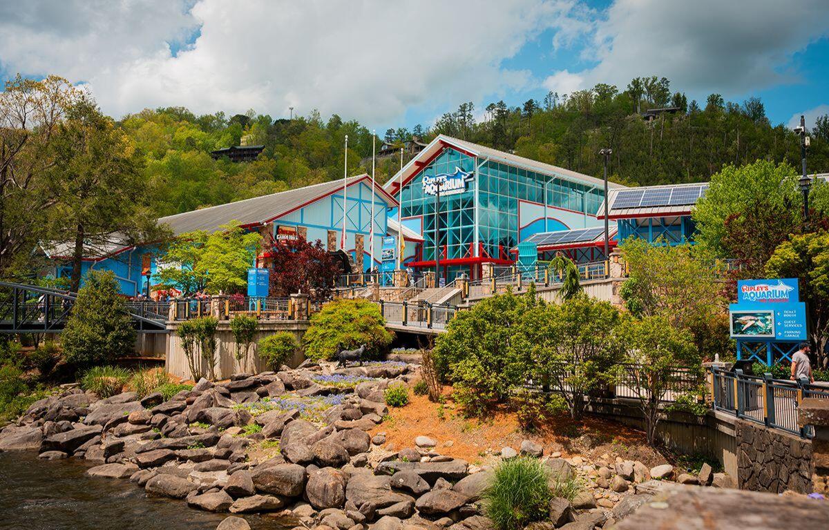 Ripley's Aquarium with blue and red building, glass facade, walkway, and rocky shoreline by water amid trees.