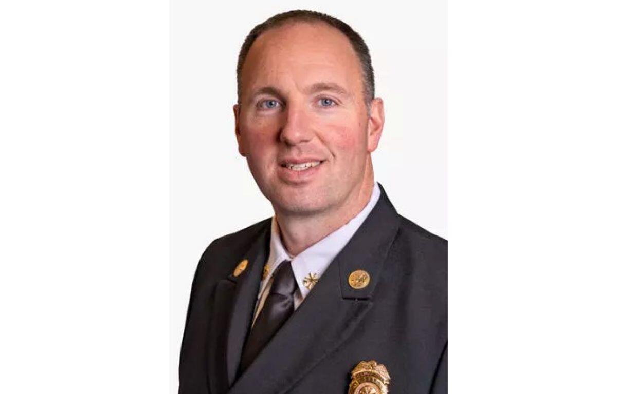 Portrait of a man in a dark suit and tie, wearing gold lapel pins and a badge, smiling at the camera.