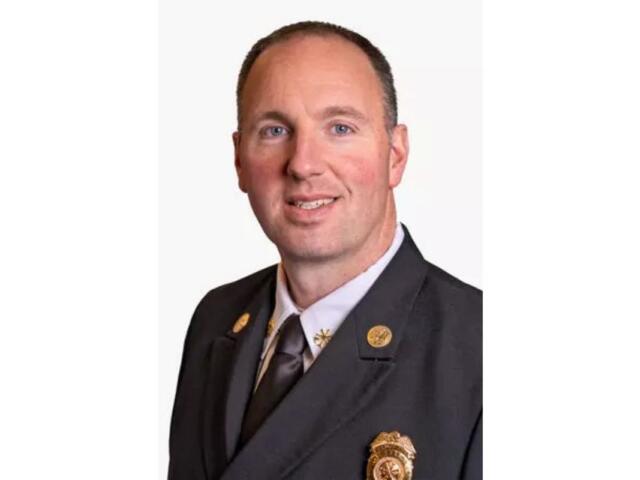 Portrait of a man in a dark suit and tie, wearing gold lapel pins and a badge, smiling at the camera.