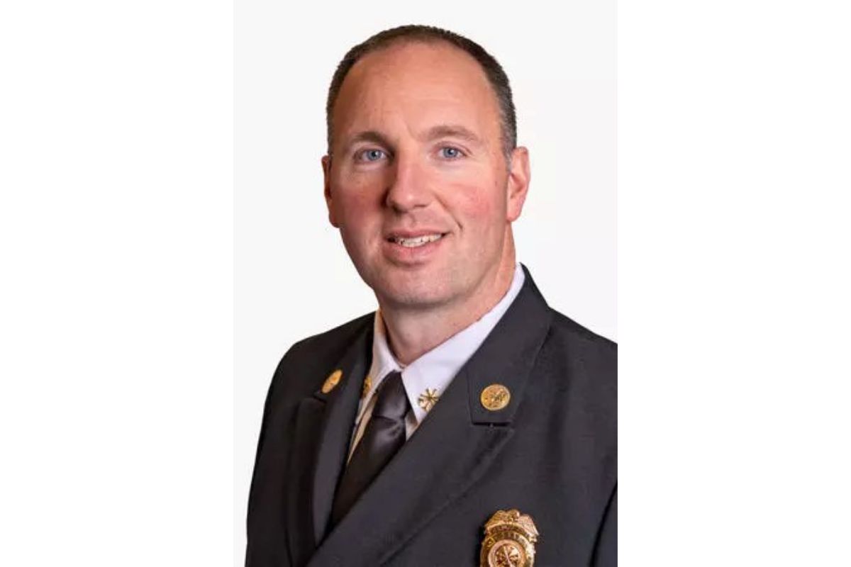 Portrait of a man in a dark suit and tie, wearing gold lapel pins and a badge, smiling at the camera.
