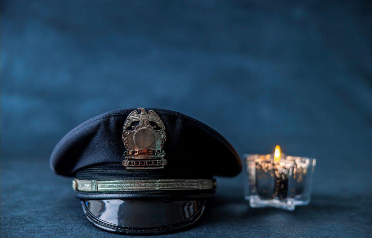 Police hat with brass badge resting on a surface beside a lit candle against a dark blue background.