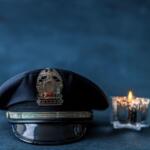Police hat with brass badge resting on a surface beside a lit candle against a dark blue background.