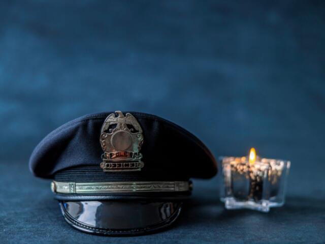 Police hat with brass badge resting on a surface beside a lit candle against a dark blue background.