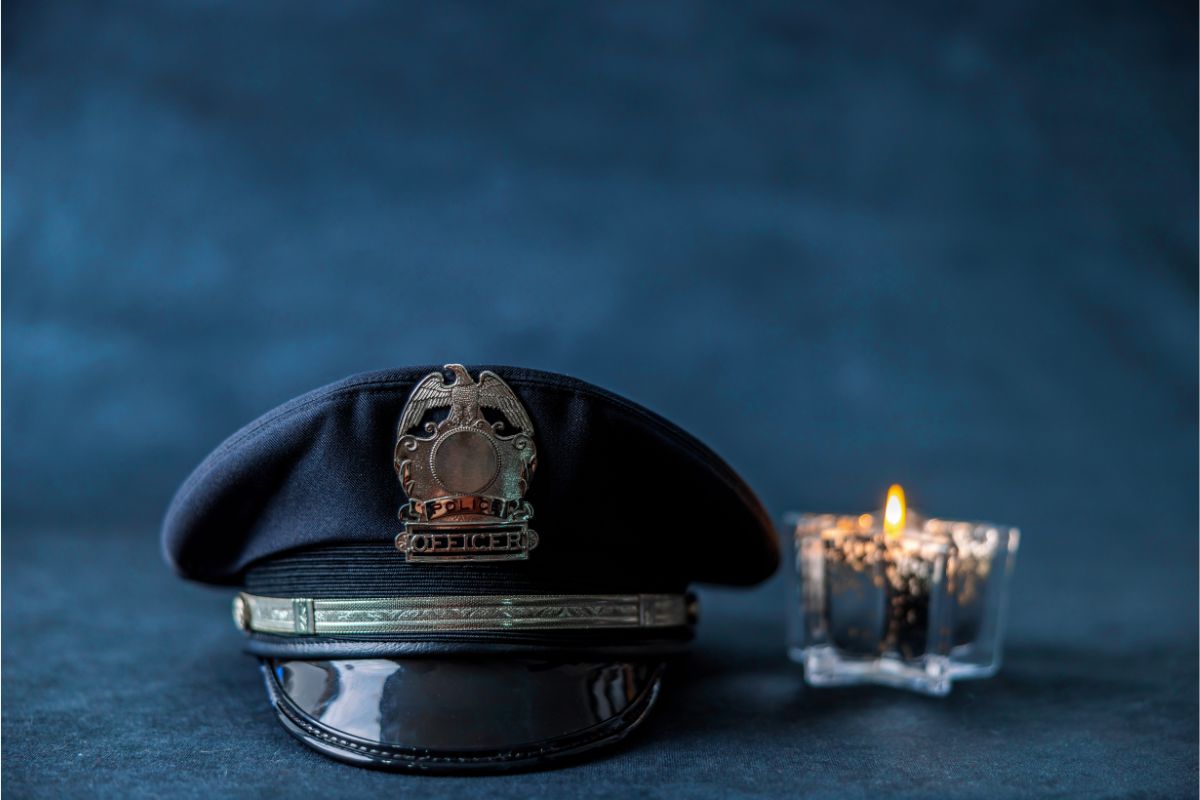 Police hat with brass badge resting on a surface beside a lit candle against a dark blue background.