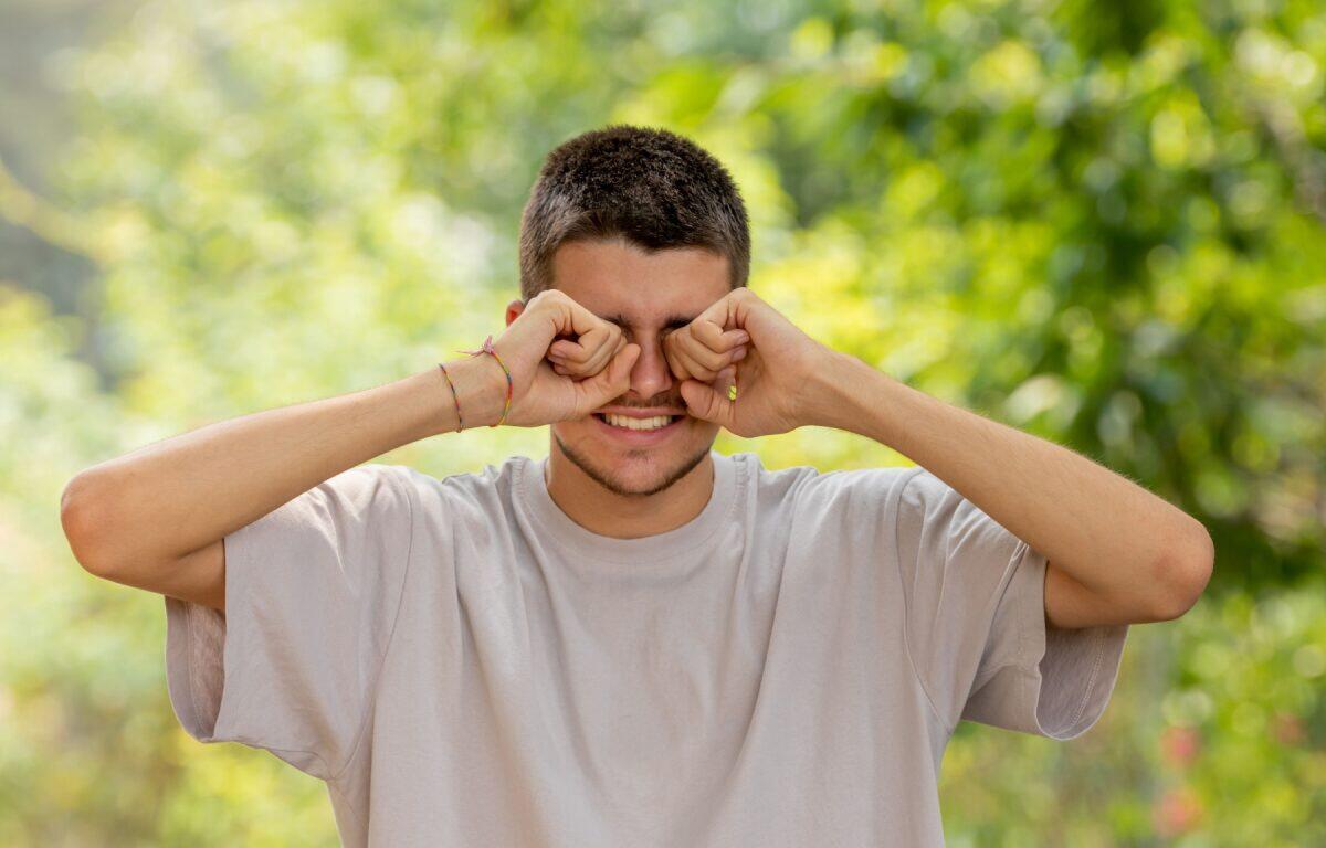 Man outdoors in a light gray t-shirt rubs his eyes with fists, smiling, in a sunny green park.