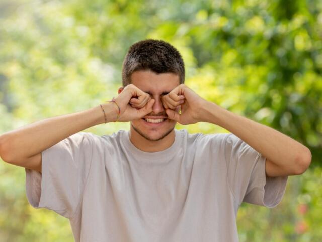 Man outdoors in a light gray t-shirt rubs his eyes with fists, smiling, in a sunny green park.