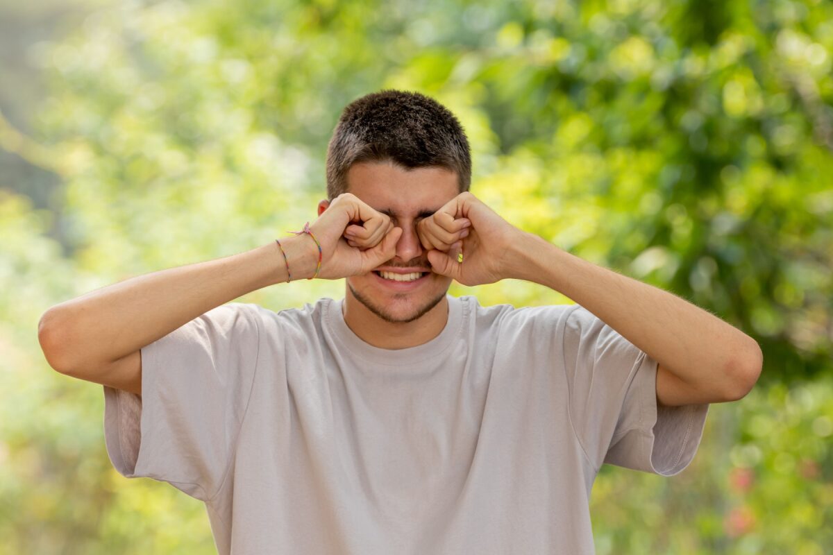 Man outdoors in a light gray t-shirt rubs his eyes with fists, smiling, in a sunny green park.