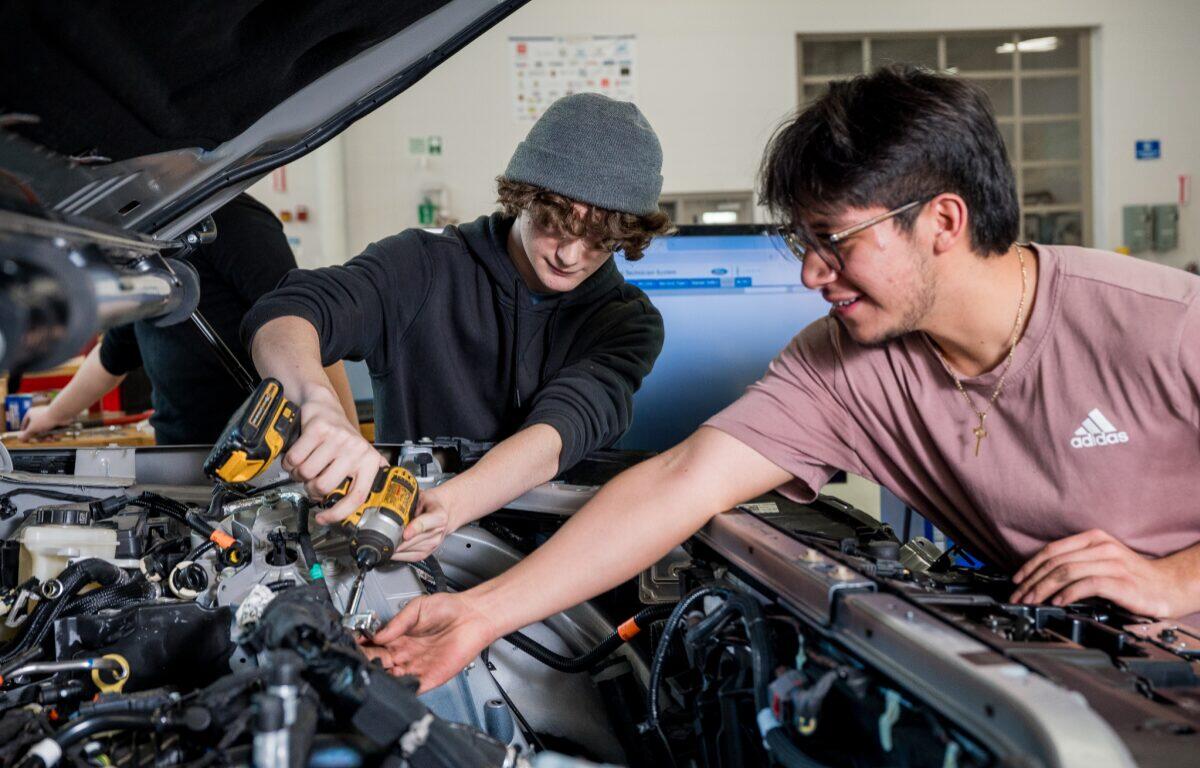 Two young men work under an open car hood; one uses a cordless drill while the other points toward the engine.