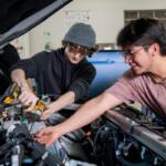 Two young men work under an open car hood; one uses a cordless drill while the other points toward the engine.