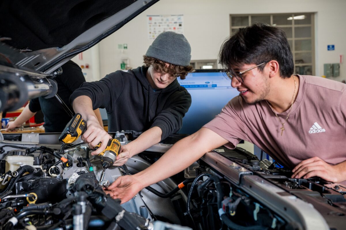 Two young men work under an open car hood; one uses a cordless drill while the other points toward the engine.