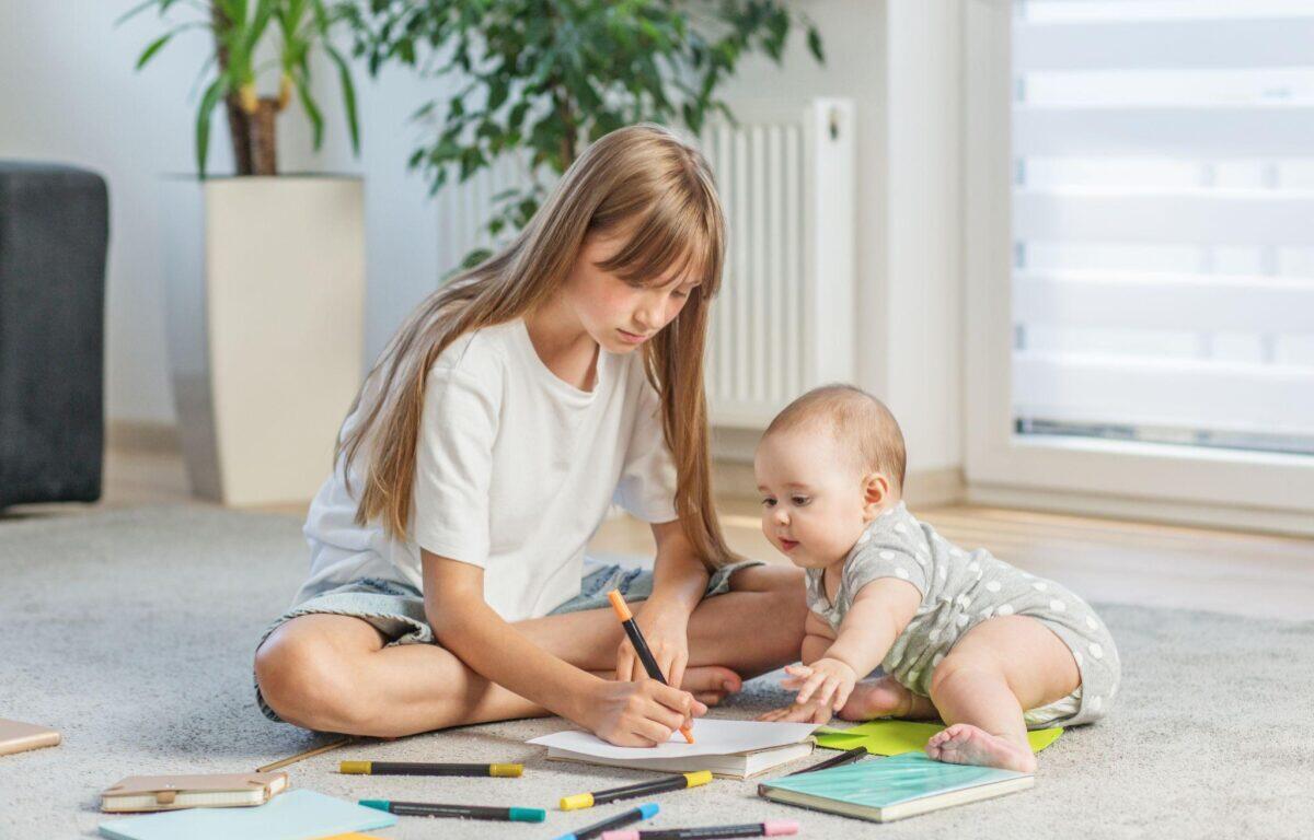 Older girl drawing with an orange marker on paper while a baby watches, surrounded by colored pencils and notebooks on the living room carpet.