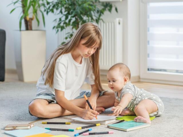 Older girl drawing with an orange marker on paper while a baby watches, surrounded by colored pencils and notebooks on the living room carpet.