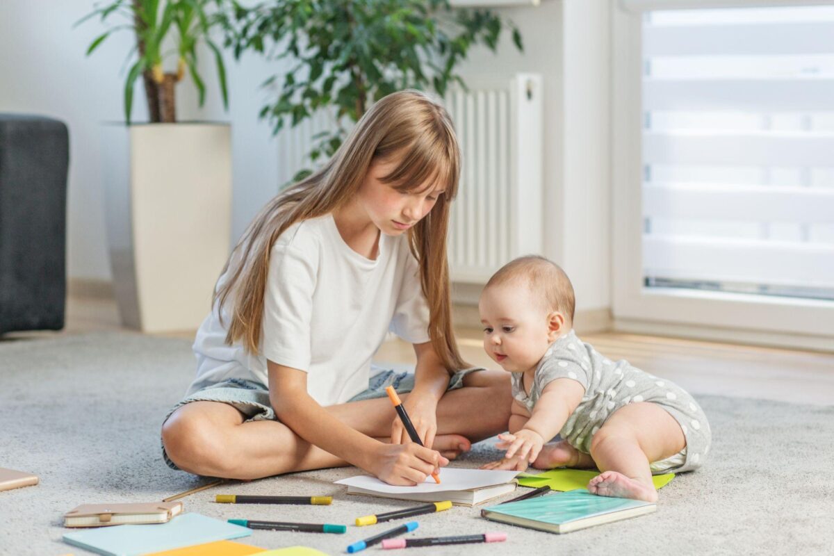 Older girl drawing with an orange marker on paper while a baby watches, surrounded by colored pencils and notebooks on the living room carpet.