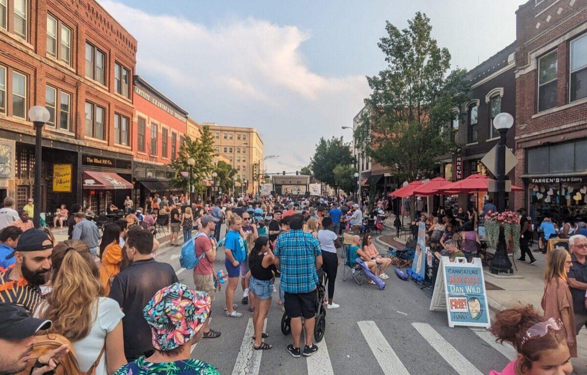 Crowded street festival with people dining under red umbrellas along a brick storefront-lined street.