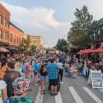 Crowded street festival with people dining under red umbrellas along a brick storefront-lined street.