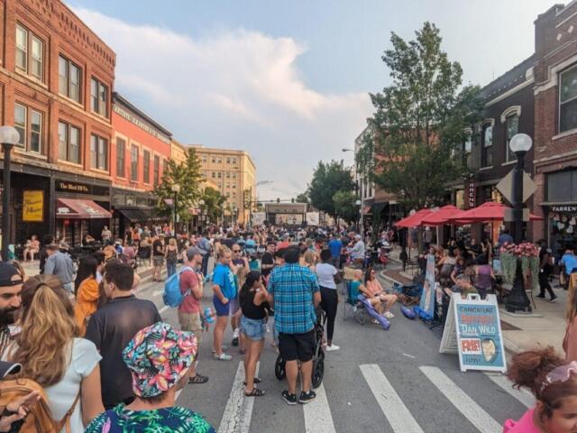 Crowded street festival with people dining under red umbrellas along a brick storefront-lined street.