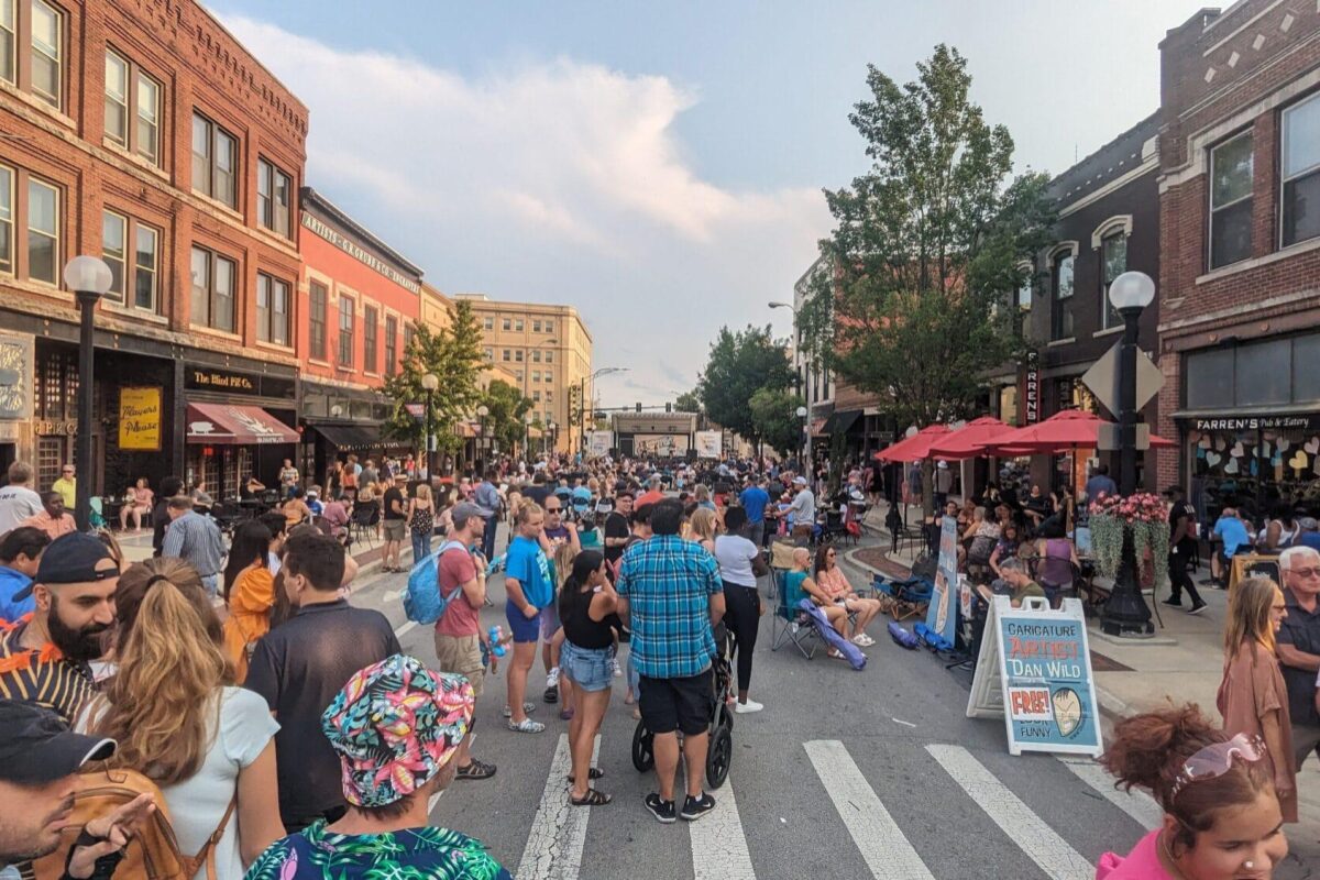 Crowded street festival with people dining under red umbrellas along a brick storefront-lined street.