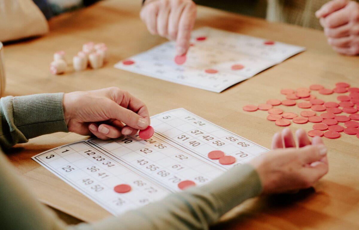 Two people play bingo, placing red chips on numbered cards at a wooden table (close-up).