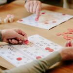 Two people play bingo, placing red chips on numbered cards at a wooden table (close-up).