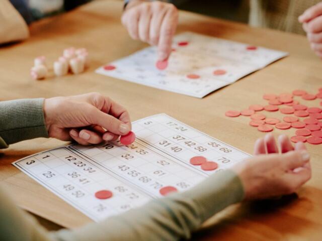 Two people play bingo, placing red chips on numbered cards at a wooden table (close-up).
