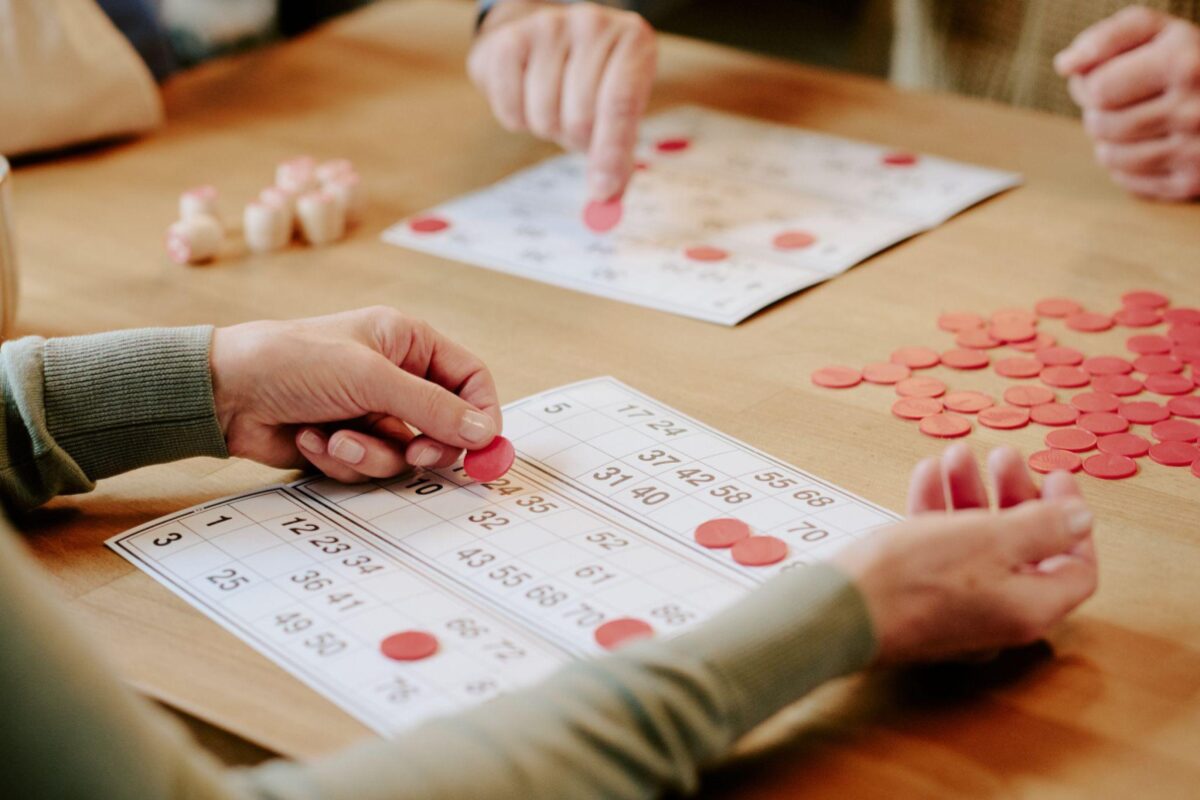 Two people play bingo, placing red chips on numbered cards at a wooden table (close-up).