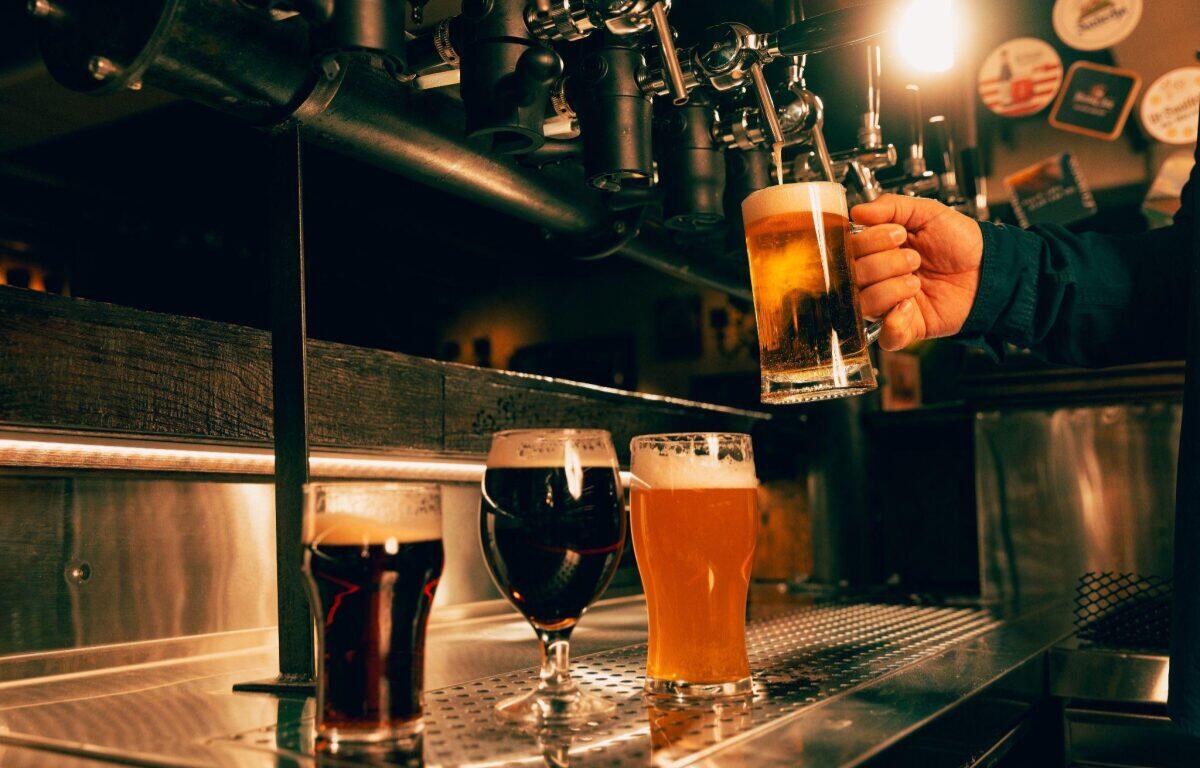Bartender pours beer from a tap into a tall glass while two other beers sit on the stainless bar nearby.