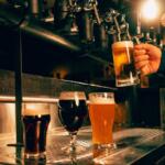 Bartender pours beer from a tap into a tall glass while two other beers sit on the stainless bar nearby.