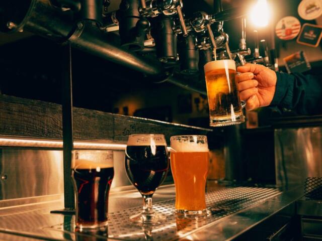 Bartender pours beer from a tap into a tall glass while two other beers sit on the stainless bar nearby.