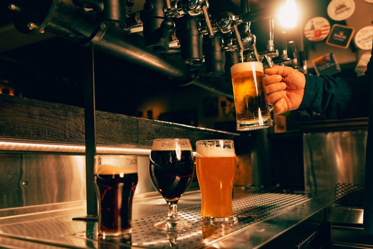 Bartender pours beer from a tap into a tall glass while two other beers sit on the stainless bar nearby.