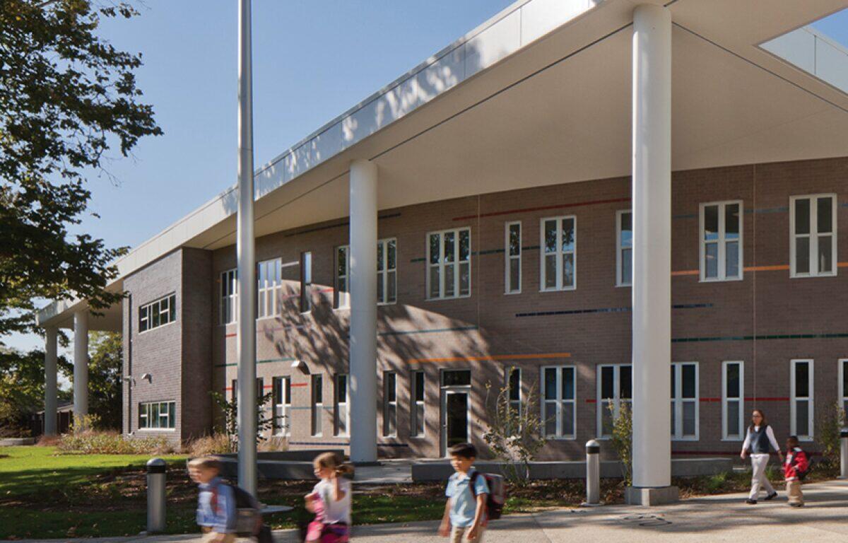 School building with large white columns and students walking along the walkway outside a brick facade building on a sunny day.