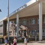 School building with large white columns and students walking along the walkway outside a brick facade building on a sunny day.
