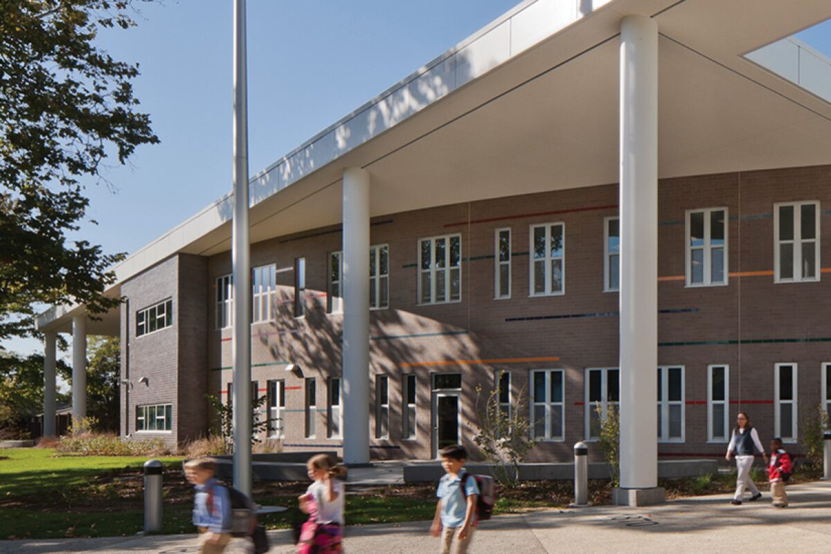 School building with large white columns and students walking along the walkway outside a brick facade building on a sunny day.