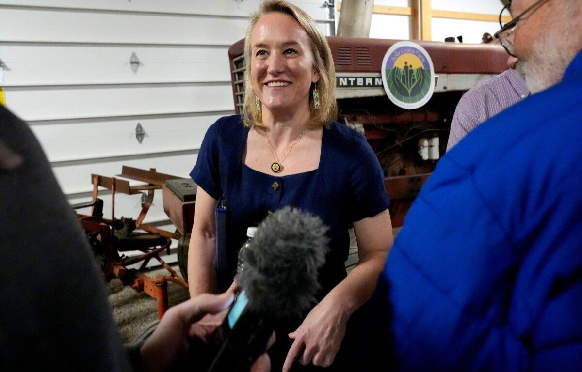 Woman in a navy dress smiles as a reporter with a microphone interviews her in a workshop with farm equipment in the background.