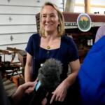 Woman in a navy dress smiles as a reporter with a microphone interviews her in a workshop with farm equipment in the background.