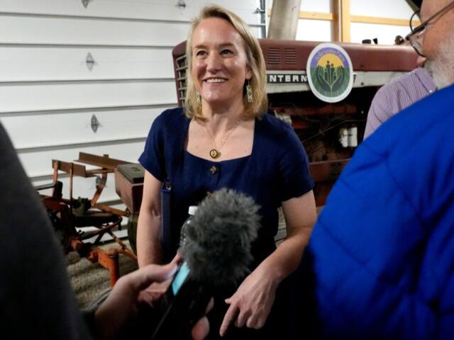 Woman in a navy dress smiles as a reporter with a microphone interviews her in a workshop with farm equipment in the background.