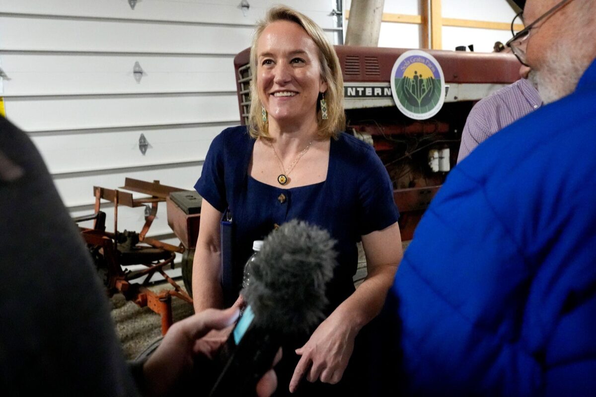 Woman in a navy dress smiles as a reporter with a microphone interviews her in a workshop with farm equipment in the background.